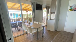 Een moderne, goed verlichte kamer met een houten vloer, een eethoek met tafel en stoelen, een bank, een aan de muur bevestigde televisie en een schuifdeur naar een terras.