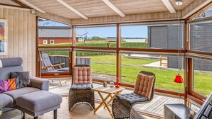 Una habitación amplia con techo de madera, ventanas grandes, un sofá gris, sillas de mimbre y una pequeña mesa.
