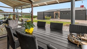 Un área de comedor exterior moderna con terraza de madera, mobiliario de mimbre negro y un recinto de vidrio con vistas a la propiedad.