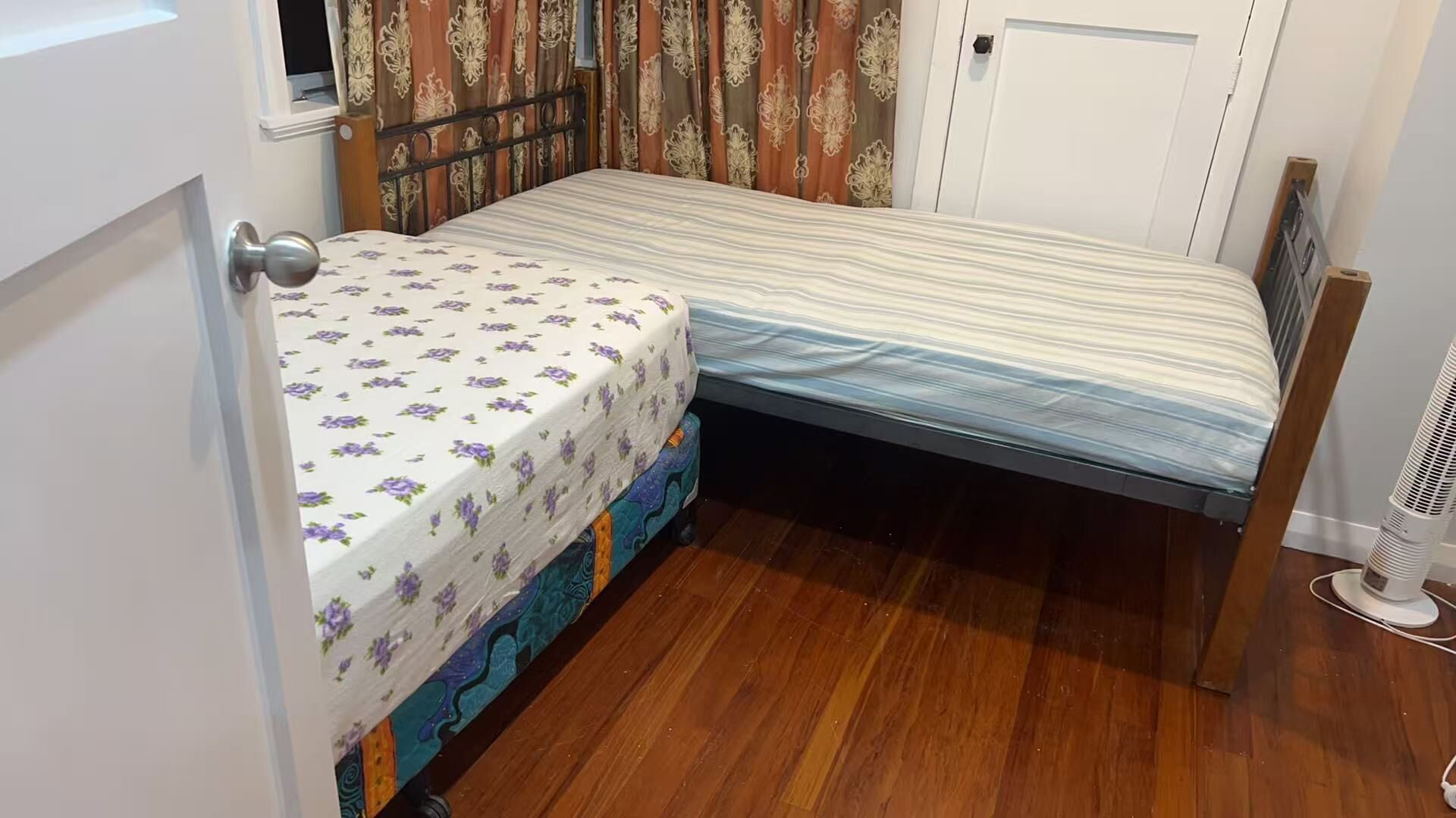 Una habitación pequeña y funcional con una cama individual, una ventana con cortinas, una puerta y un ventilador de pie.