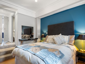 Ein Schlafzimmer mit einem großen Bett, einem Schreibtisch und einem Stuhl. Eine Tür führt in einen weiteren Raum.