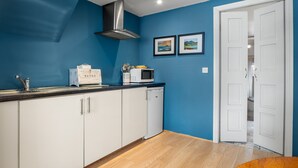 A compact kitchen with white cabinets, a stainless steel range hood, a microwave, and a bread box on the counter. There are two framed pictures on the wall and a white door leading to another room.