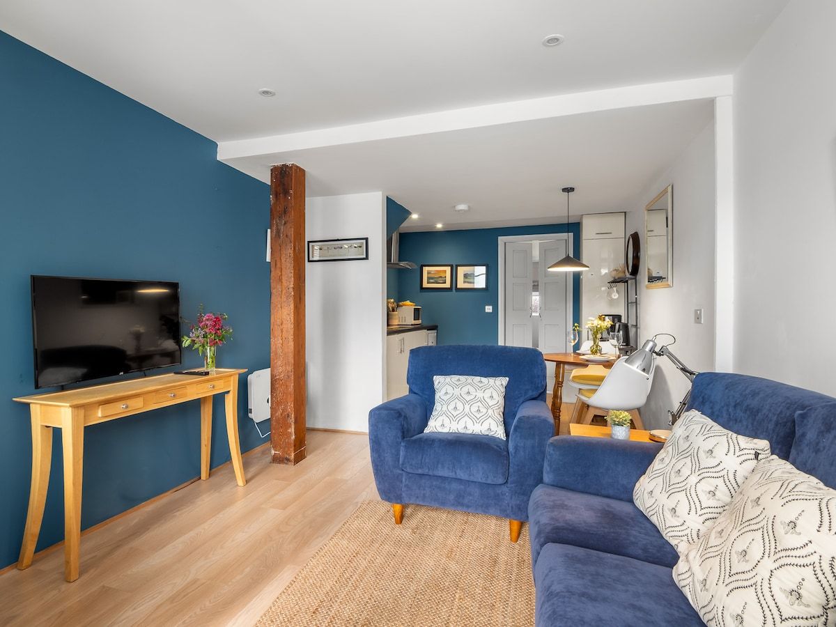 A modern living room with a television, wooden desk, blue sofa, and a dining area in the background.
