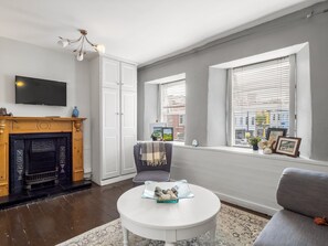 A living room with a fireplace, a television, a round coffee table, and a window with blinds. - Clifden Nook 2 Apt w/ Fireplace (Clifden)