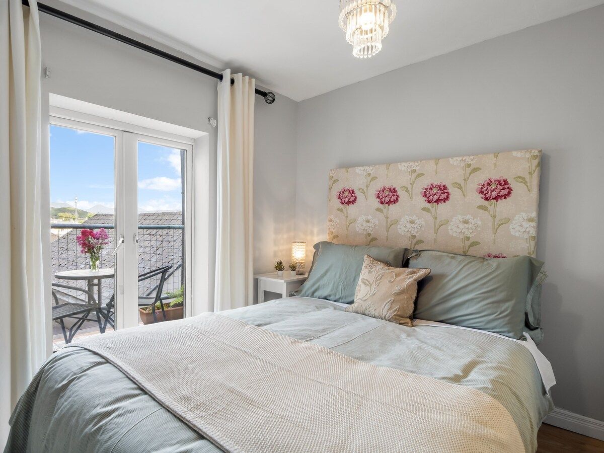 A bedroom with a bed, a nightstand, a lamp, and a view of the outside.