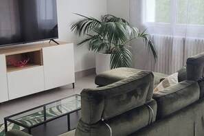 Un soggiorno moderno con un divano verde, una TV a schermo piatto su un supporto in legno, una pianta in vaso e un tavolino da caffè in vetro.