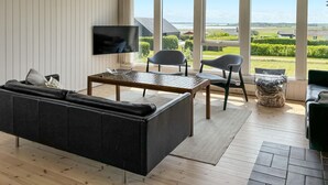 A modern living room with a large window, a flat-screen TV, a dark sofa, two chairs, and a coffee table. - 3 bedroom stunning home in øster assels (Øster Assels)