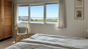 A bedroom with a large bed, a wooden chair, a window with a view of houses and a body of water, and a white wall with a framed picture. - 3 bedroom stunning home in øster assels (Øster Assels)