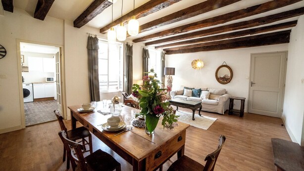 A dining area with a wooden table set for four, a sofa in the background, and exposed wooden beams on the ceiling.