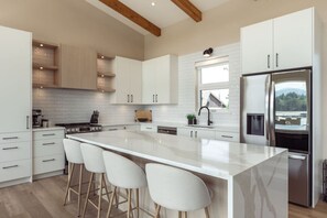 A modern kitchen with white cabinetry, a large island with bar stools, a stainless steel refrigerator, and a window providing a view.