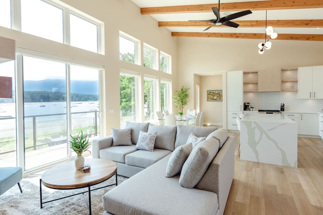 Un salon spacieux avec un canapé gris, une petite table ronde et une vue sur un lac, visible à travers de grandes fenêtres.