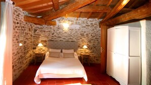 Un dormitorio con pared de piedra, techo de madera, cama, lámparas de noche y un armario.