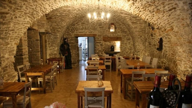 Interior de paredes de piedra con mesas y sillas de madera, un candelabro y botellas de vino sobre una mesa.