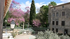 Un patio con un edificio de piedra, un árbol alto y árboles en floración de color rosa.