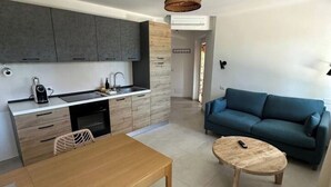 A modern kitchen with grey cabinets, a wooden dining table, and a blue sofa in the living area. - Agreen Country House Juniper (Sardegna)
