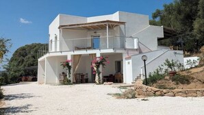A modern two-story house with a covered outdoor area, a stone retaining wall, and a gravel driveway. - Agreen Country House Juniper (Sardegna)