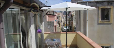 A small balcony with a table and chair, a white umbrella, and a view of the cityscape.