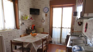 A kitchen with a dining table, chairs, a TV mounted on the wall, a clock, and a window with curtains. - Sole&Luna (Milazzo)