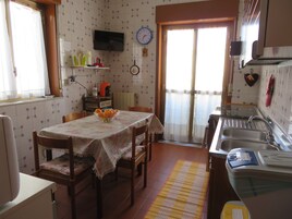 Una cocina con una mesa de comedor, sillas, un televisor montado en la pared, un reloj y una ventana con cortinas.