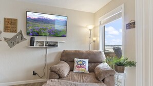 A living room with a large flat-screen TV, a comfortable armchair, a small shelf with electronic devices, a wall-mounted lamp, and a window with a view of a beach. - Reel Relaxin - Waterfront Pool, Indoor Pool & Hot Tub! (Village of Four Seasons)