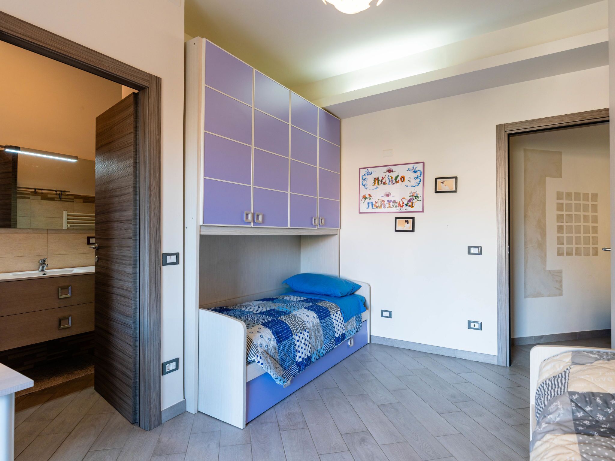 Une chambre à coucher de taille réduite, avec un lit superposé, une armoire intégrée, une petite table de chevet et une œuvre d’art accrochée au mur.