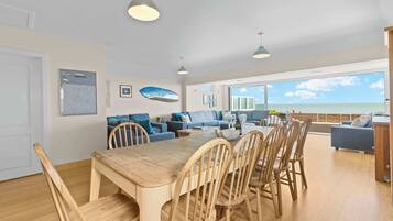 Un vaste espace de vie comprenant une table et des chaises en bois pour la salle à manger, un ensemble de canapés et une grande fenêtre donnant sur la mer.