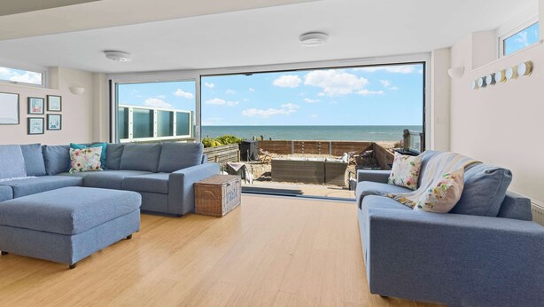 Amplio salón con un ventanal que da al mar, un conjunto de sofá gris y suelo de madera.
