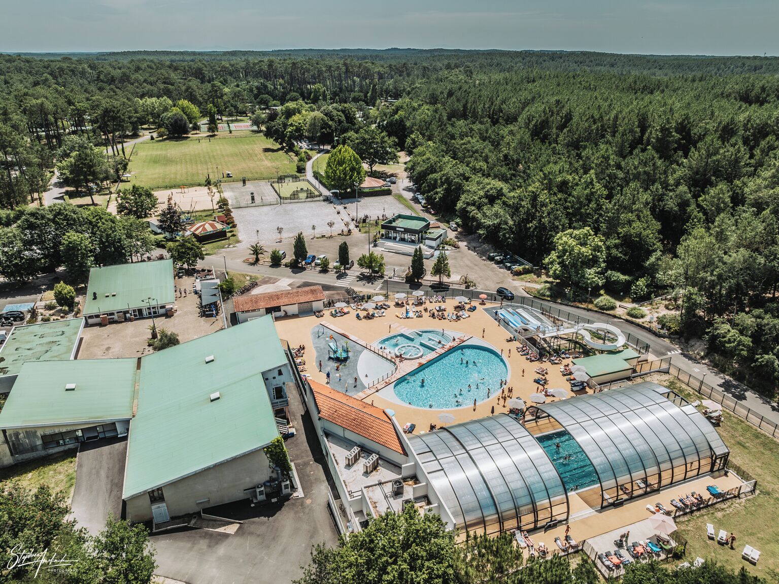 An aerial view of a resort with multiple swimming pools, a large covered area, and a surrounding forested landscape.
