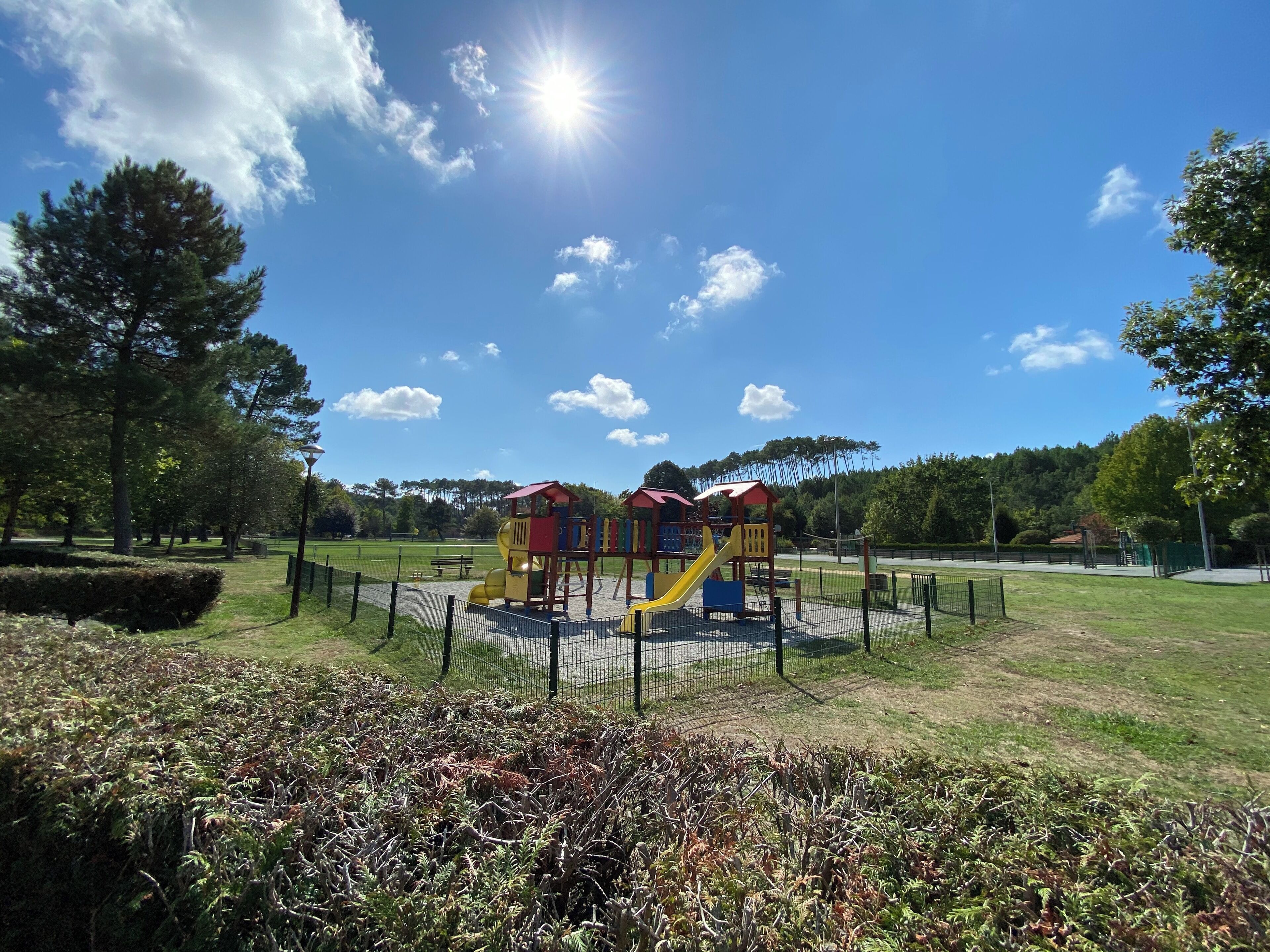 A playground with slides and climbing structures, surrounded by a fence and trees.