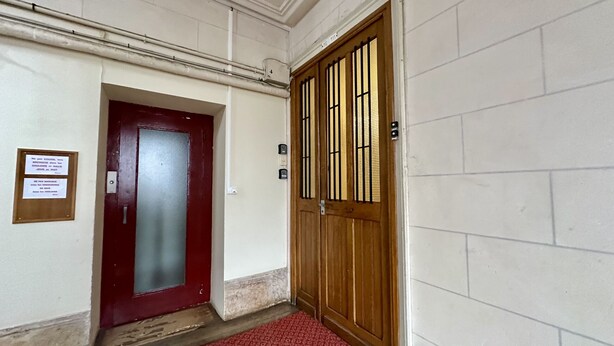 Ein Flur mit einer roten Tür und einer Holztür mit Metallgitter. An der Wand befindet sich eine Plakette und auf dem Boden ein roter Teppich.