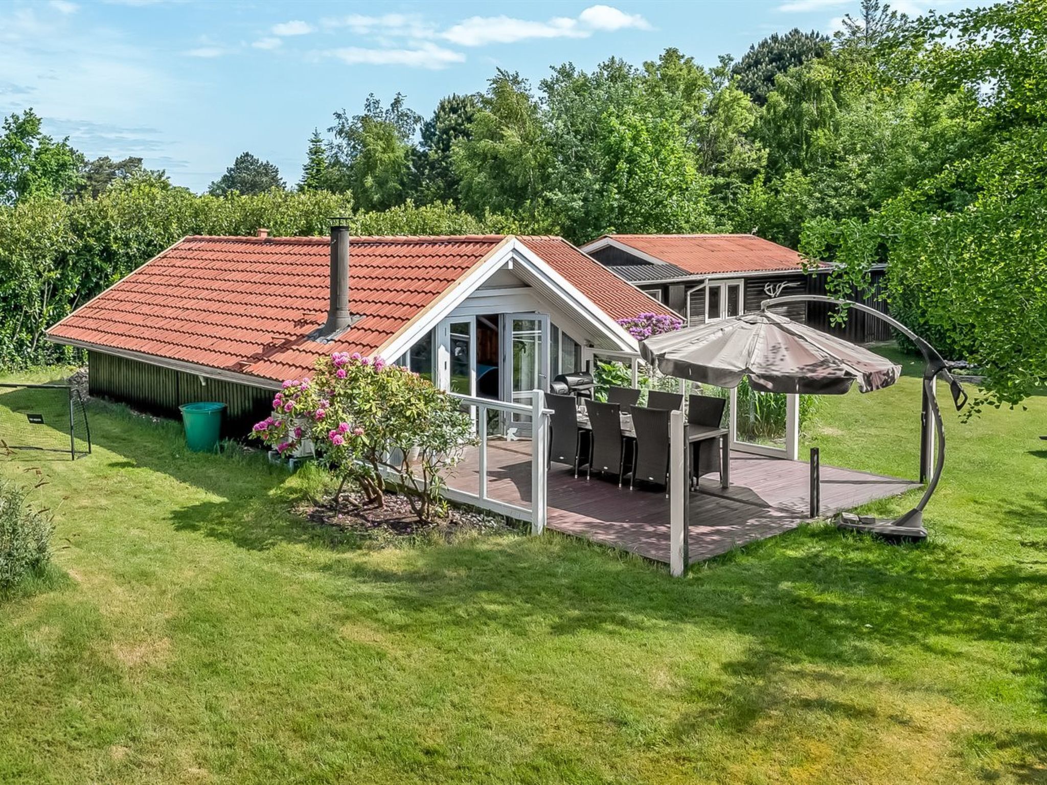 Une maison avec un toit recouvert de tuiles rouges, un patio abrité avec des meubles d’extérieur et un jardin agrémenté de plantes à fleurs.