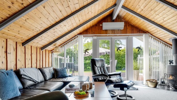 A modern loft-style room with a sloped ceiling, large windows, a black leather sofa, a dining table, and a wood-burning stove.