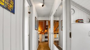 A narrow hallway with wooden floors and white walls, leading to a kitchen area with wooden cabinets and a hanging light fixture.