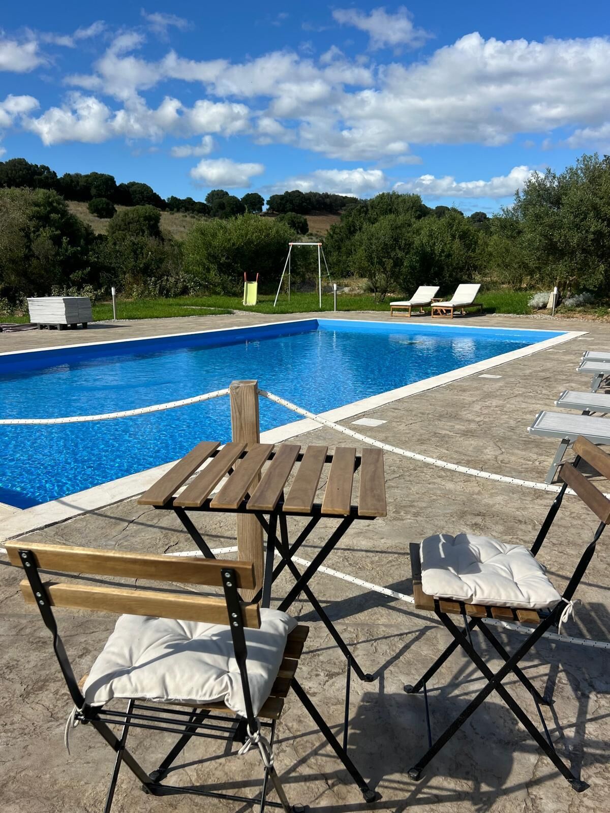 A poolside area with a wooden table and two folding chairs, a swing set, and lounge chairs in the background.