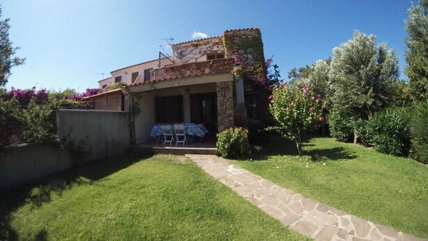 A two-story house with a stone facade, a covered patio with a table and chairs, and a well-maintained garden with various plants and flowers. - Villa Alice very close to the sea (Sardegna)