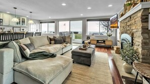 A spacious living room with a large sofa, a coffee table, and a fireplace. There is a kitchen area with cabinets and a dining table. - Lakefront 5 Bds, sleeps 8 -Blue Waters Cottage (Ransomville)