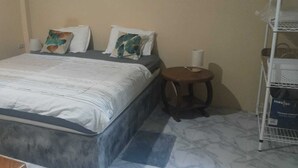 A bed with a grey headboard, a small round wooden stool, and a white vacuum cleaner on a shelf.