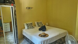 A single bed with a white bedspread, two patterned pillows, a rolled-up towel, and a straw hat on the bed. There is a standing fan to the right of the bed.
