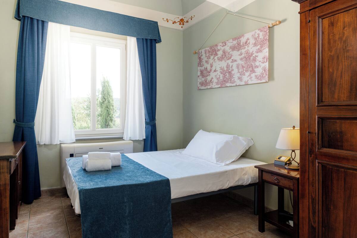 Ein Schlafzimmer mit einem Einzelbett, einem Nachttisch mit Lampe, einem Fenster mit Vorhängen und einem an der Wand befestigten Kunstwerk.