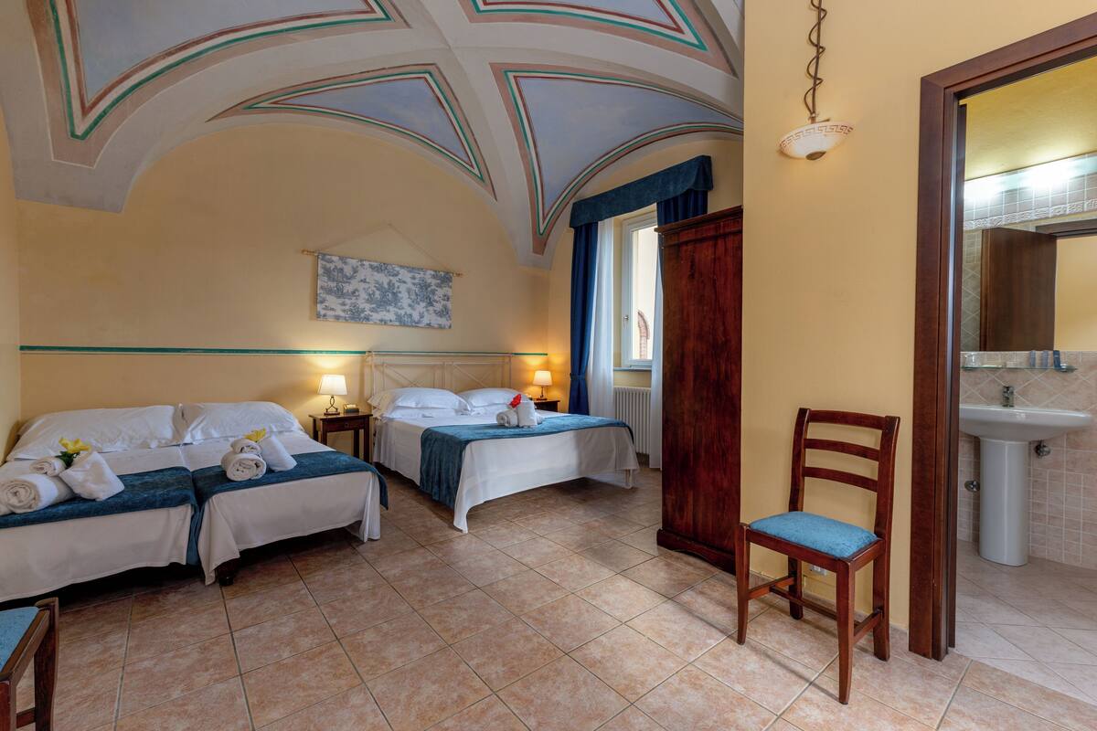 Ein Hotelzimmer mit zwei Betten, einem Holzhocker, einem Badezimmer mit Waschbecken und einer dekorativen Decke.