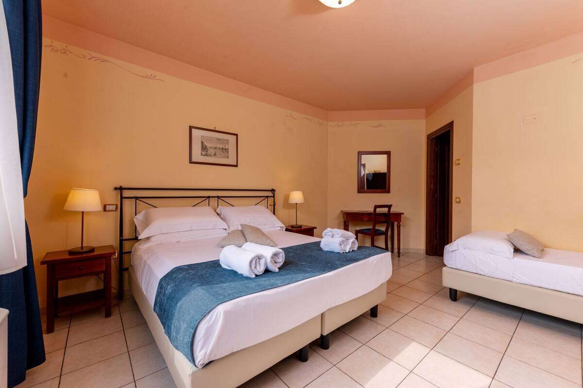 Ein Hotelzimmer mit einem großen Bett, einem kleineren Bett, zwei Nachttischen, einem Schreibtisch, einem Stuhl, einem Spiegel und einem gerahmten Bild an der Wand.