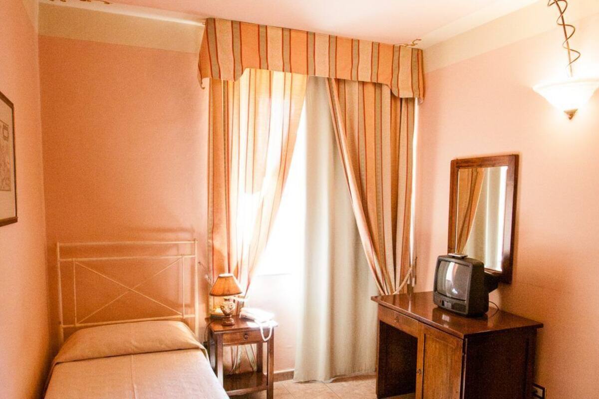 Ein Hotelzimmer mit einem Bett, einem Nachttisch mit Lampe, einem Fernseher und Vorhängen.