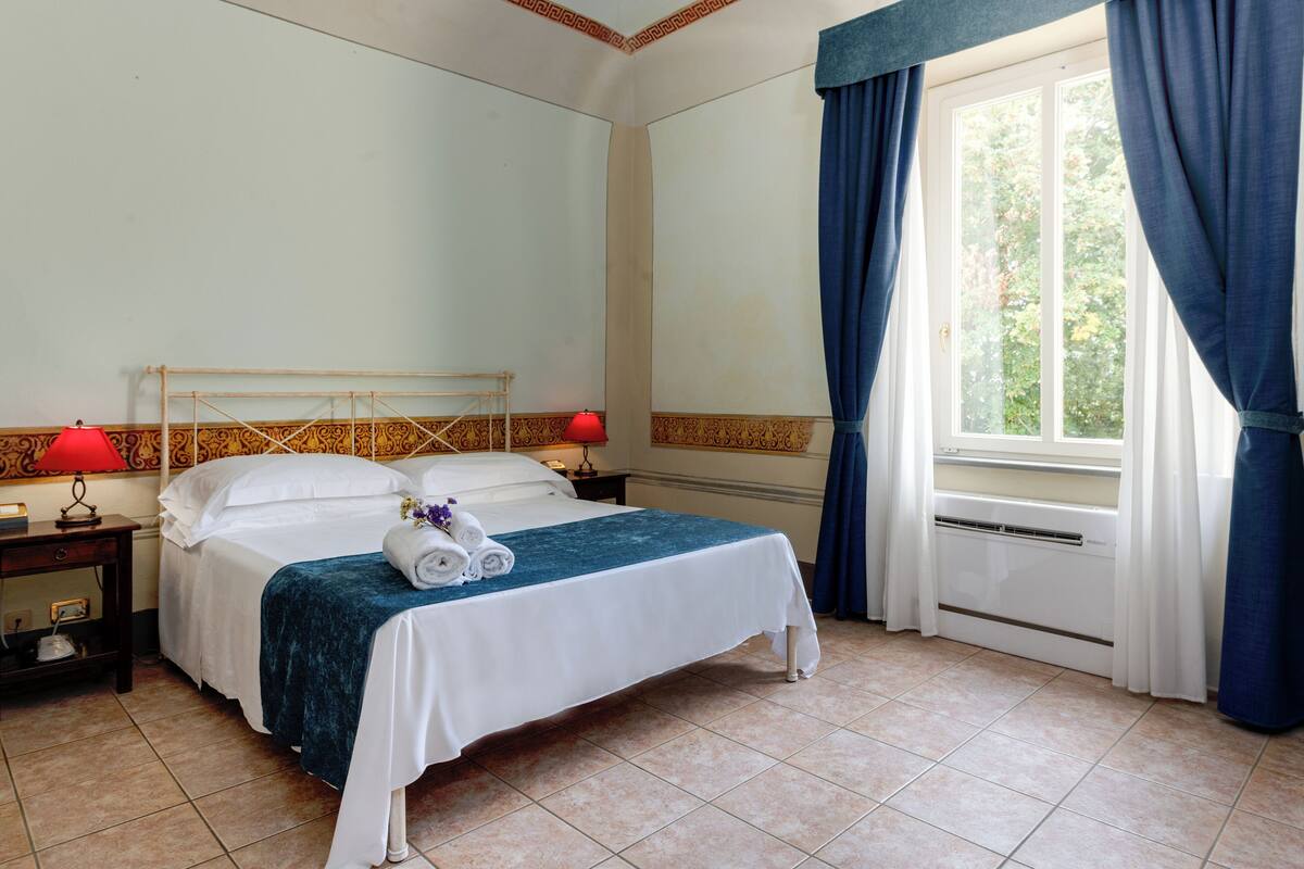 Ein ordentlich bezogenes Bett mit blauer Tagesdecke, weißen Kissen und zusammengerollten Handtüchern. Ein Nachttisch mit roter Lampe und ein kleiner Tisch mit einer Lampe stehen bereit. Ein Fenster mit blauen Vorhängen ist sichtbar, und ein Heizkörper befindet sich auf dem Boden.