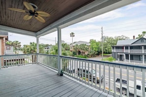 A covered porch with a ceiling fan, white railing, and a view of a street with parked cars and houses.