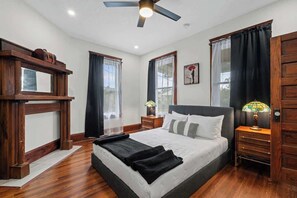 A bedroom with a large bed, wooden headboard, two bedside tables with lamps, a ceiling fan, and a window with curtains.