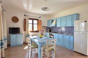 Une cuisine avec des meubles de couleur bleu clair, un réfrigérateur blanc, une table à manger avec des chaises, un téléviseur posé sur un meuble et une fenêtre avec des rideaux.