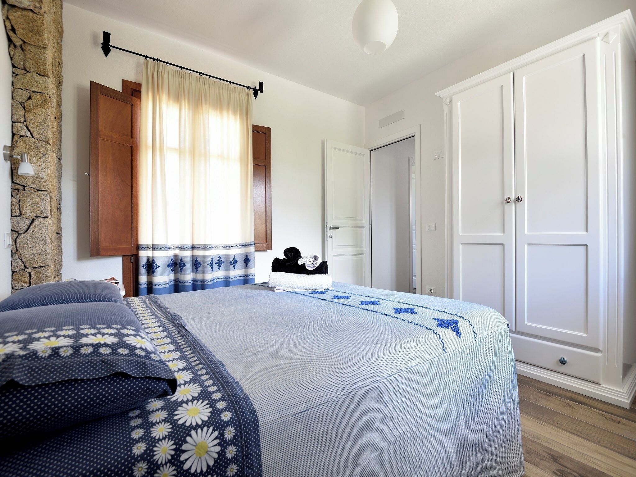 Une chambre à coucher avec un lit, une armoire, un mur en pierre et une fenêtre avec des rideaux.