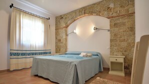 A bedroom with a bed, a nightstand, a chair, a window with curtains, and a stone wall. - Villa Menhir! Exclusive property close to the beach (Lotzorai)