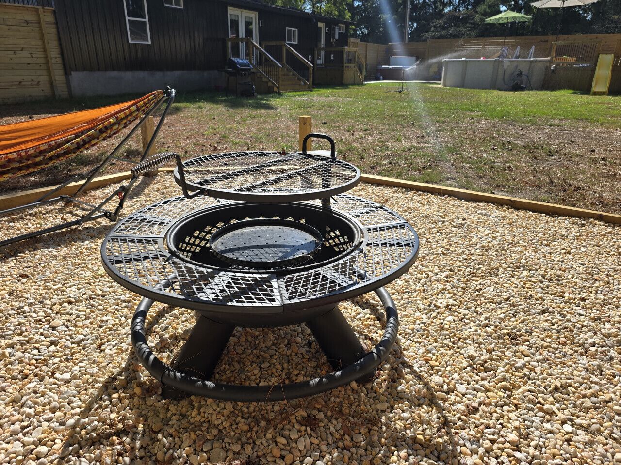 A round outdoor grill with a metal grill and a central fire pit, surrounded by gravel.