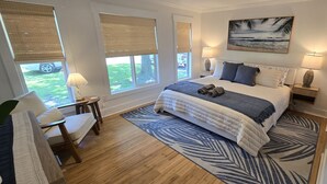 A bedroom with a bed, two bedside lamps, a large window with blinds, a chair, and a rug.